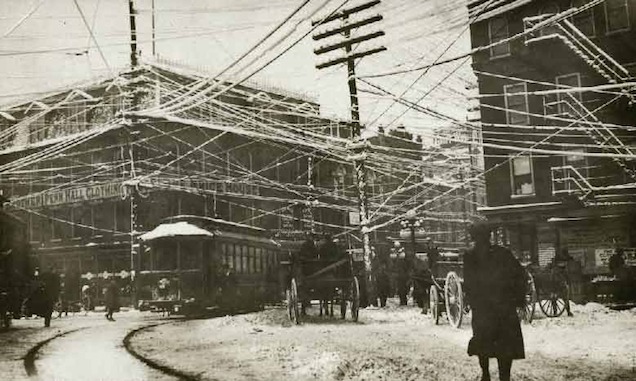 A historical black-and-white photograph showing a street scene with numerous overhead electrical wires and pedestrians, likely from the early 20th century.