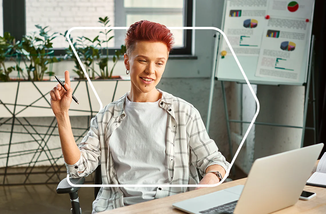 Woman with short red hair participating in an online meeting, holding a pen, with charts in the background.