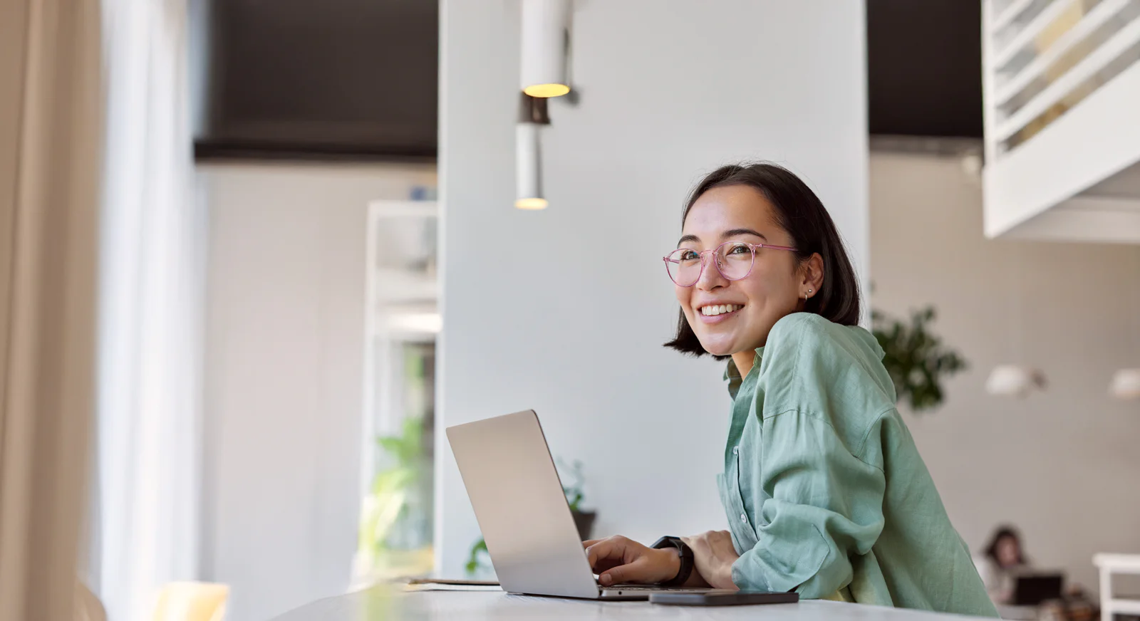 Pretty cheerful Asian woman in eyeglasses and casual clothes browses laptop computer connected to 4g internet updates software uses modern technologies poses in cafeteria looks gladfully away