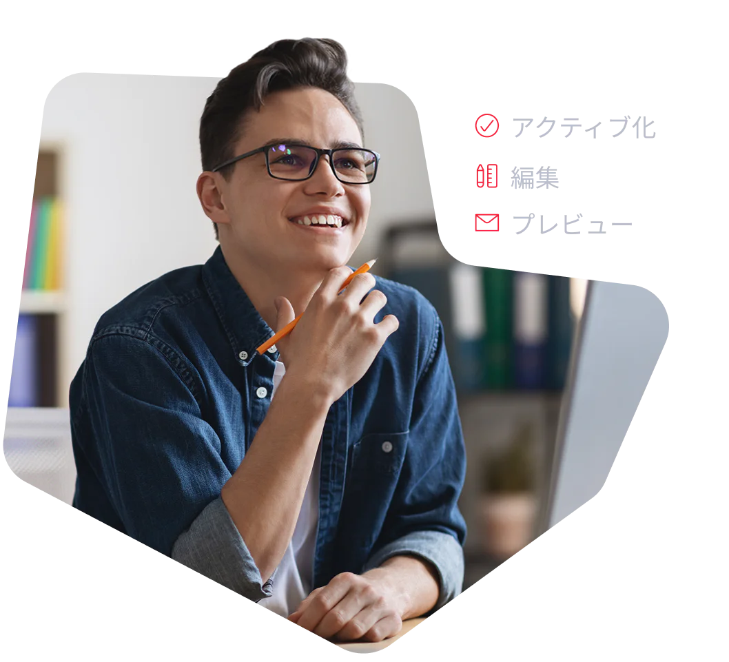 A young man with glasses and a denim shirt smiles while working at a computer.