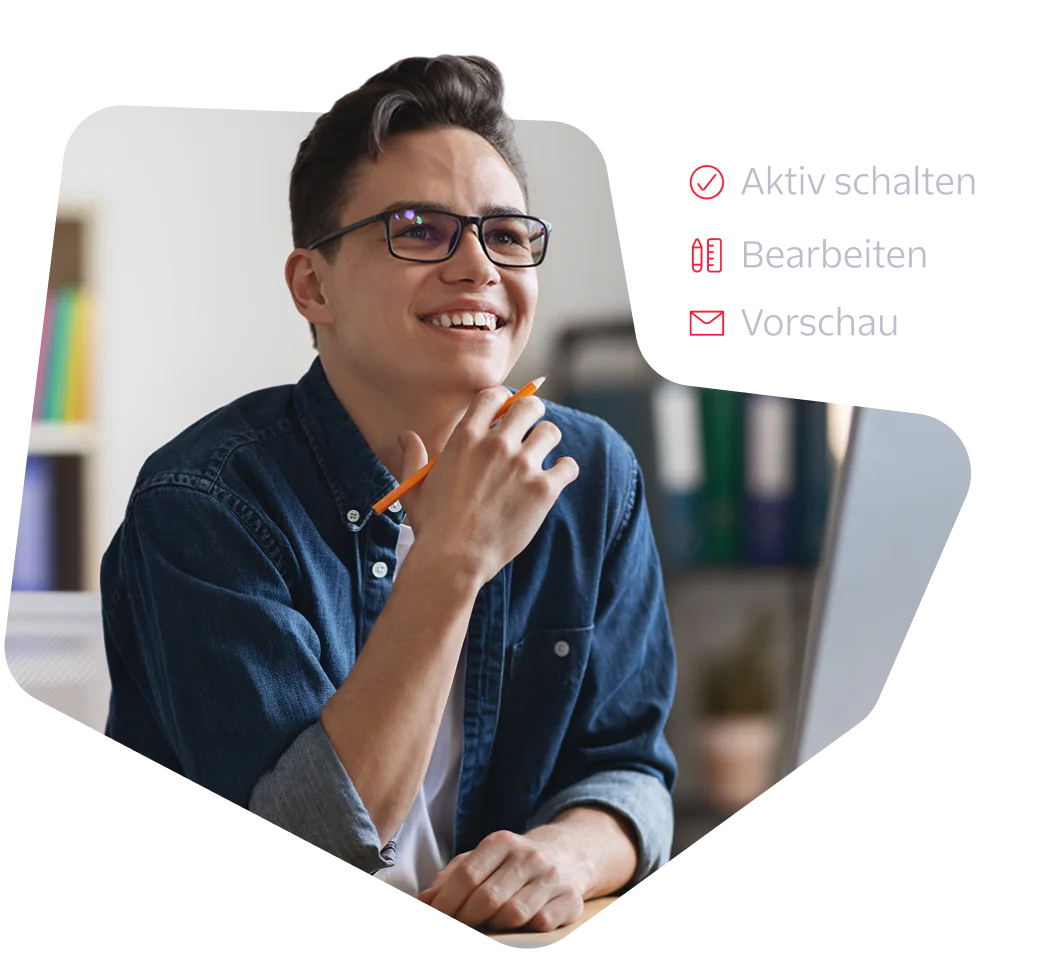 A young man with glasses and a denim shirt smiles while working at a computer.