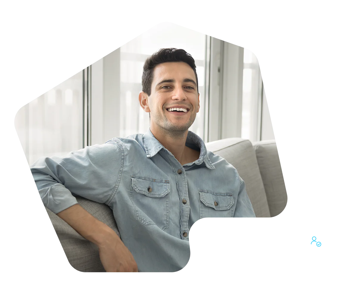 Smiling man in a light blue denim shirt sits on a gray couch in a bright room.