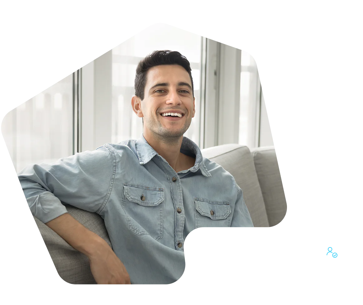 Smiling man in a light blue denim shirt sits on a gray couch in a bright room.