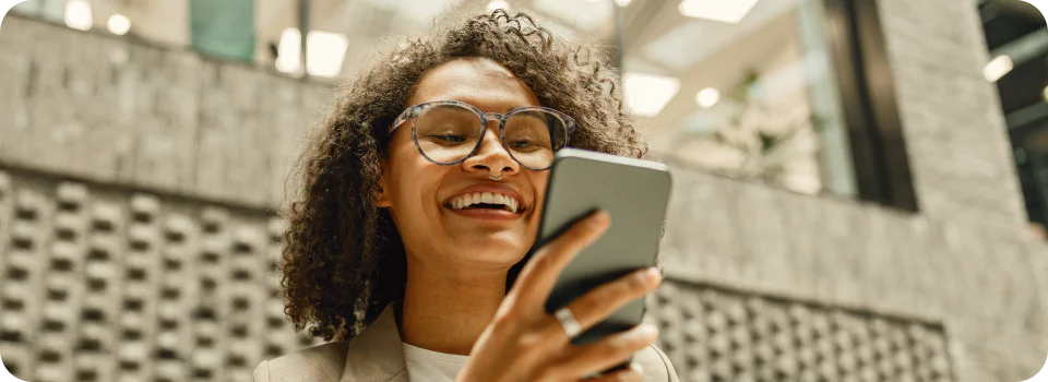 Woman with curly hair and glasses smiling at her phone outside a building.
