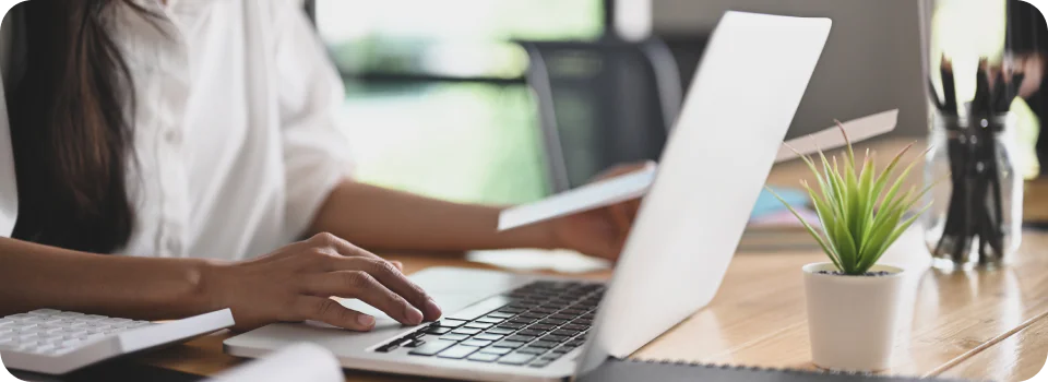 Person typing on laptop with documents, calculator, and potted plant on a wooden desk.