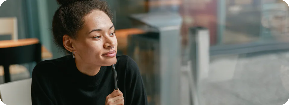 Woman holding a pen, looking thoughtfully to the side, sitting at a desk in an office environment.