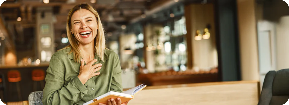 Smiling woman in green shirt holding an open book in a cozy cafe.