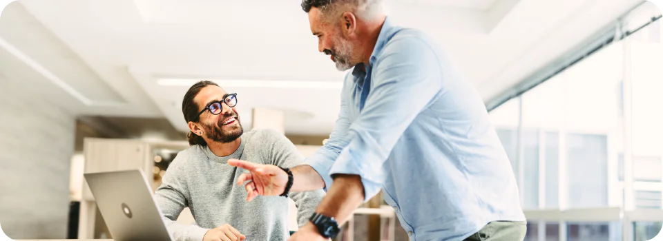Two men in discussion, one seated with a laptop, the other standing and pointing