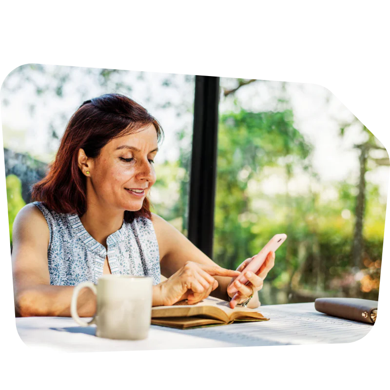 Woman reading a book, holding a phone, and drinking coffee at a table with a window view.
