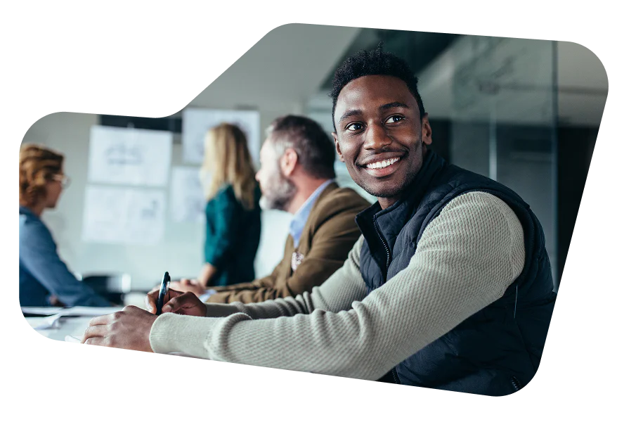 Smiling man in a vest attends a meeting with colleagues around a conference table.