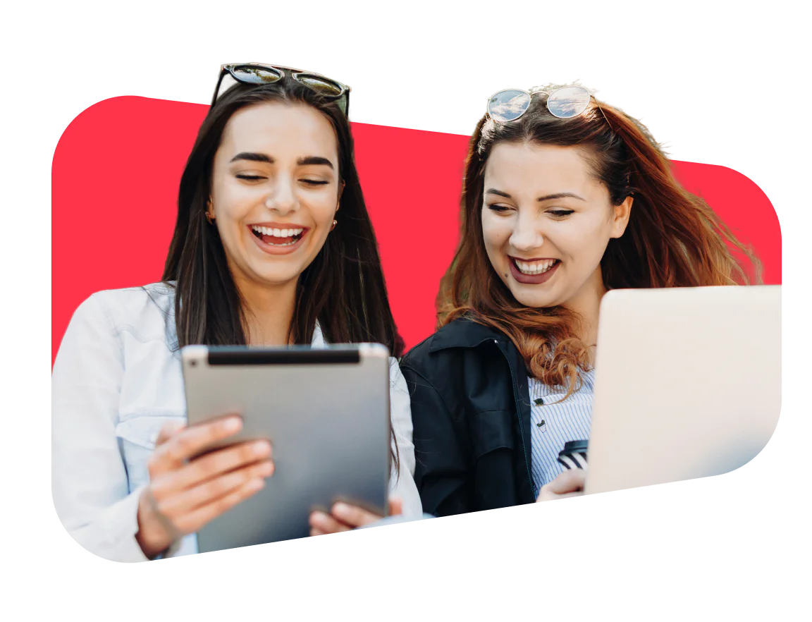Two people smiling together while using a tablet and a laptop against a pink background.