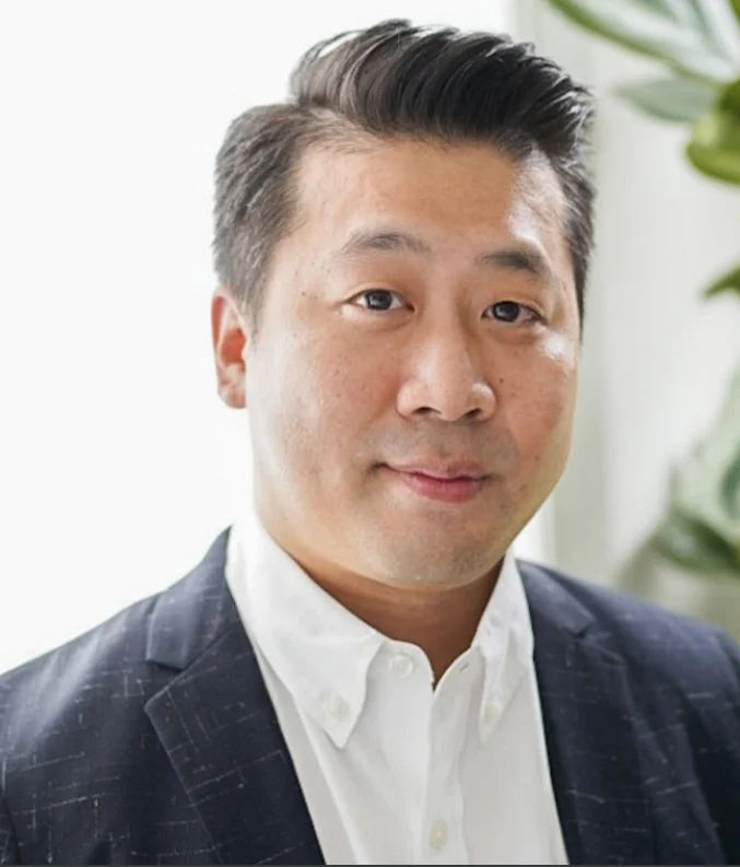 Headshot of a person in a dark blazer and white shirt, looking at the camera indoors with soft light and a plant in the background.