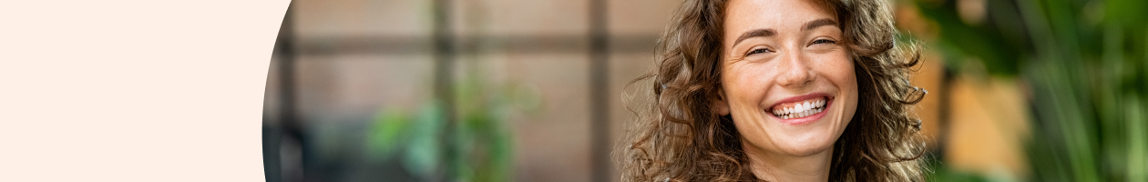 Woman with curly hair smiling in an indoor environment with greenery in the background.
