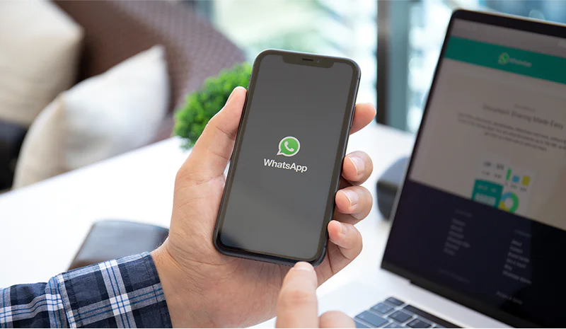 Hand holding a smartphone displaying WhatsApp logo next to an open laptop with a web page on the screen