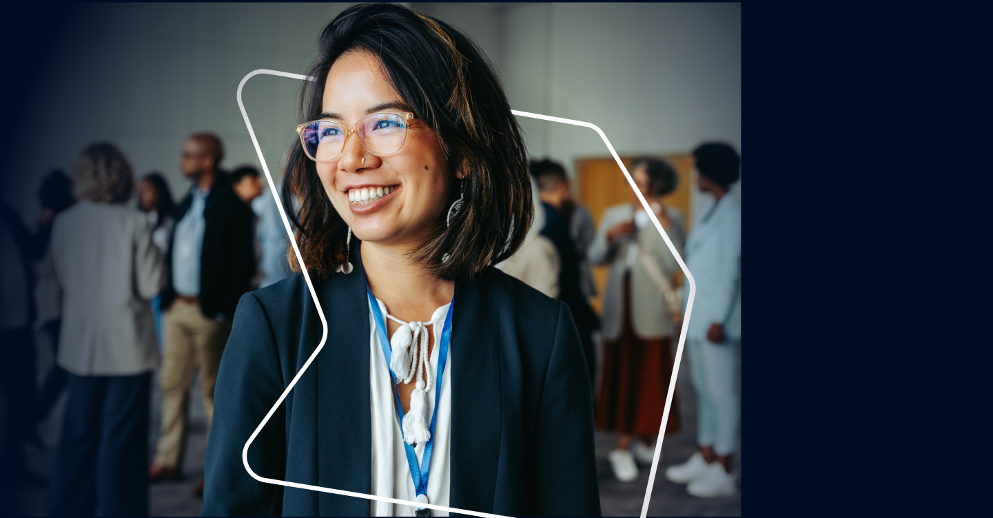 Smiling woman wearing glasses at a professional networking event with people talking in the background.