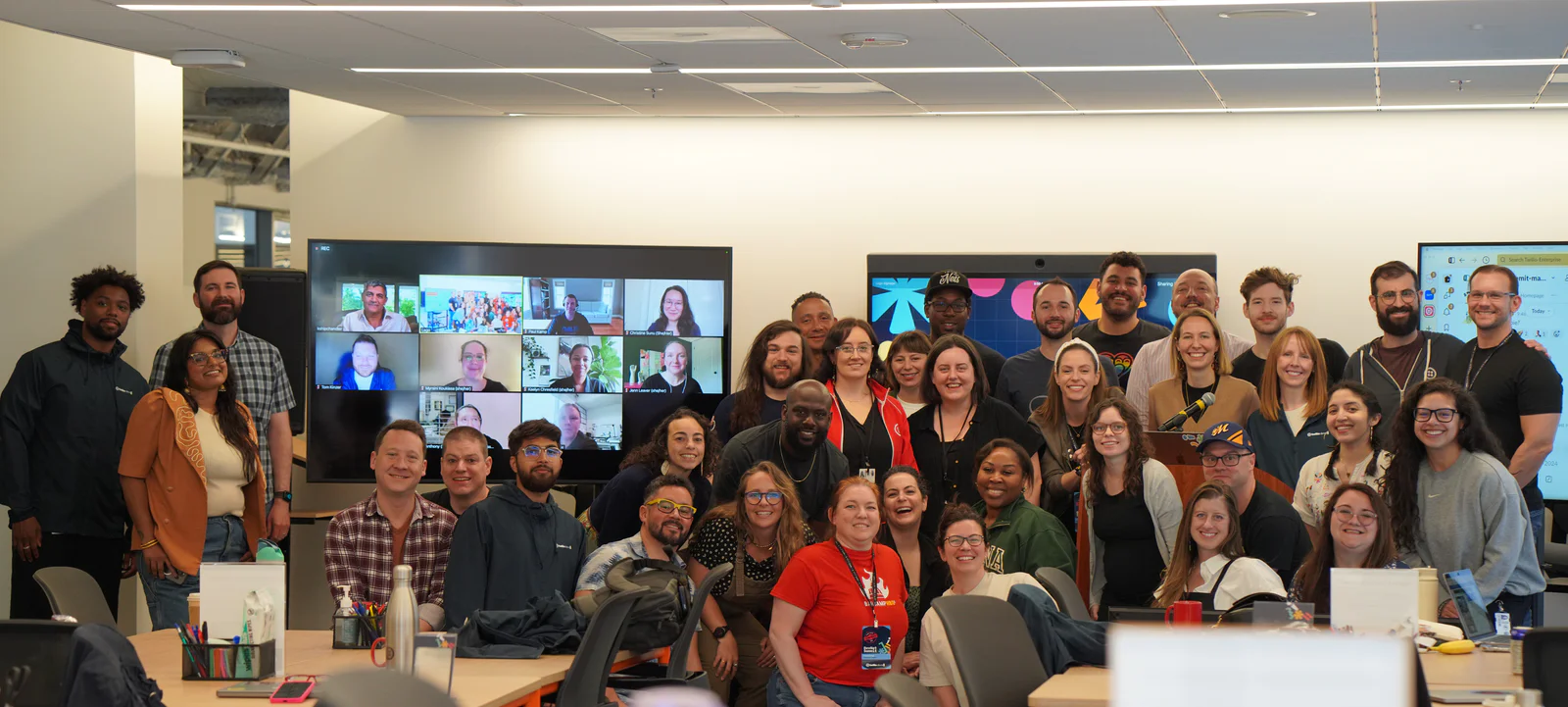 Office team posing for a group photo with some participants on video conference screens.