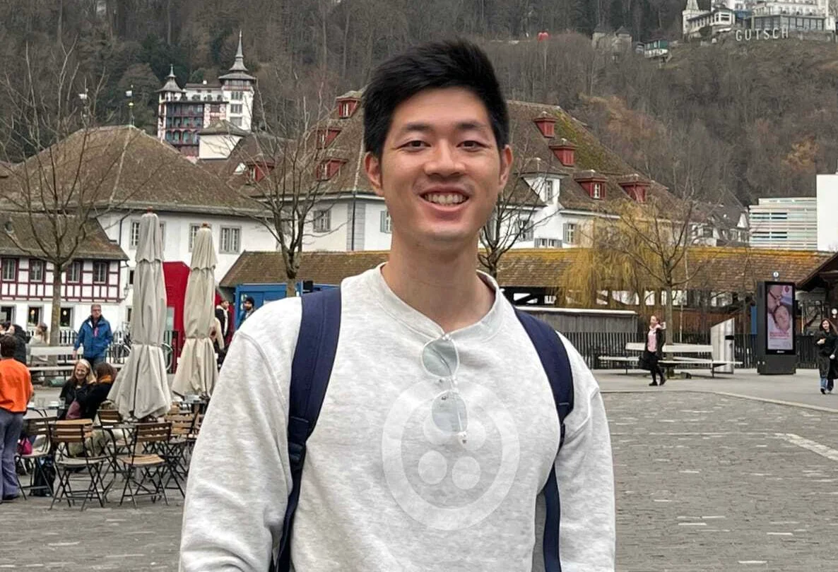 A man smiling, wearing a backpack, with European buildings and hills in the background.
