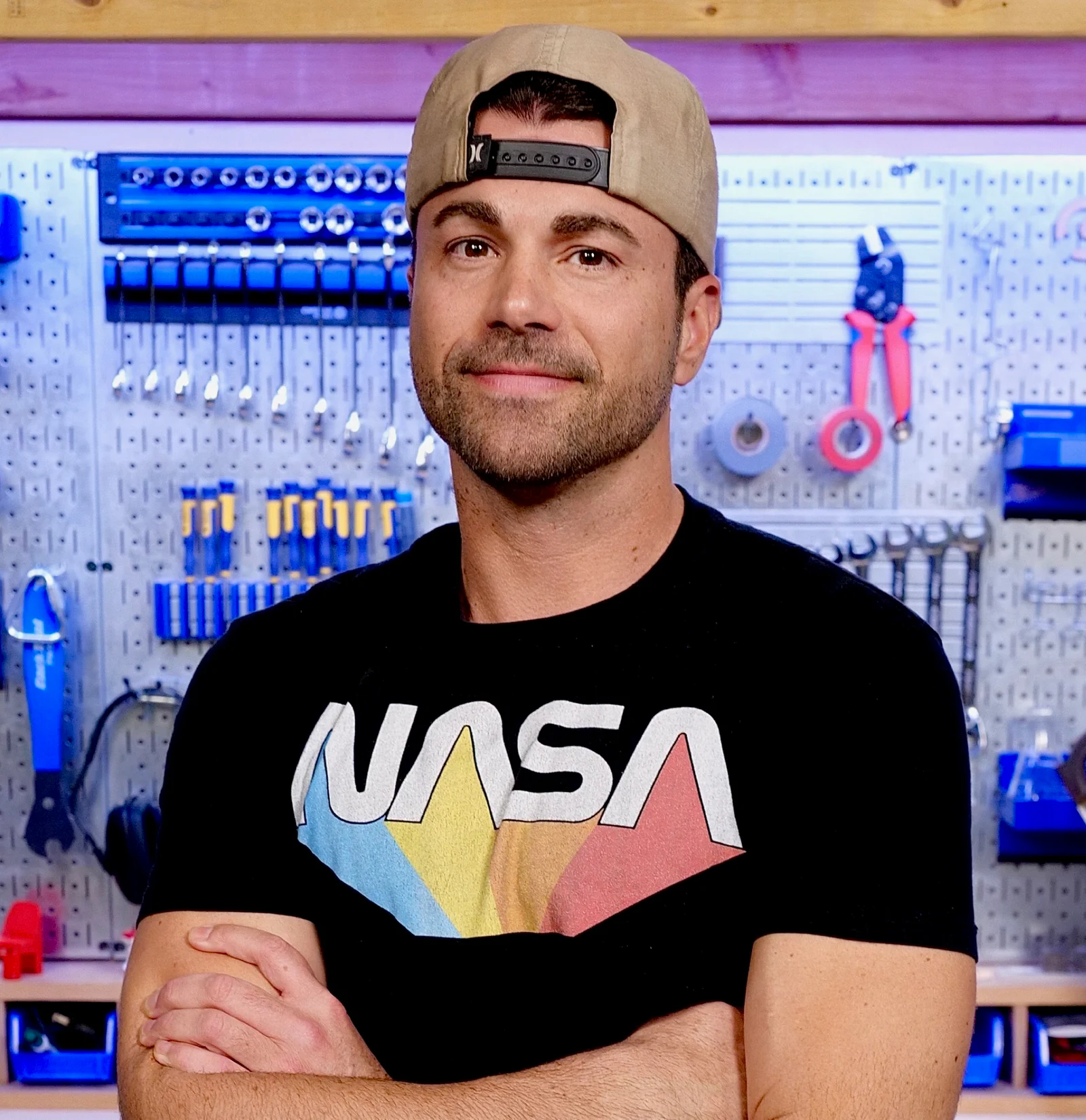 Man with a backward cap and NASA T-shirt standing with arms crossed in front of a wall of tools