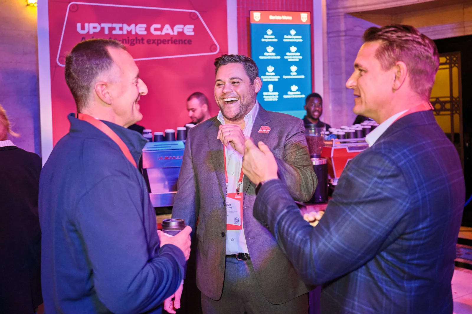 Three men in suits conversing and laughing at a networking event by a cafe stand with a sign in the background.