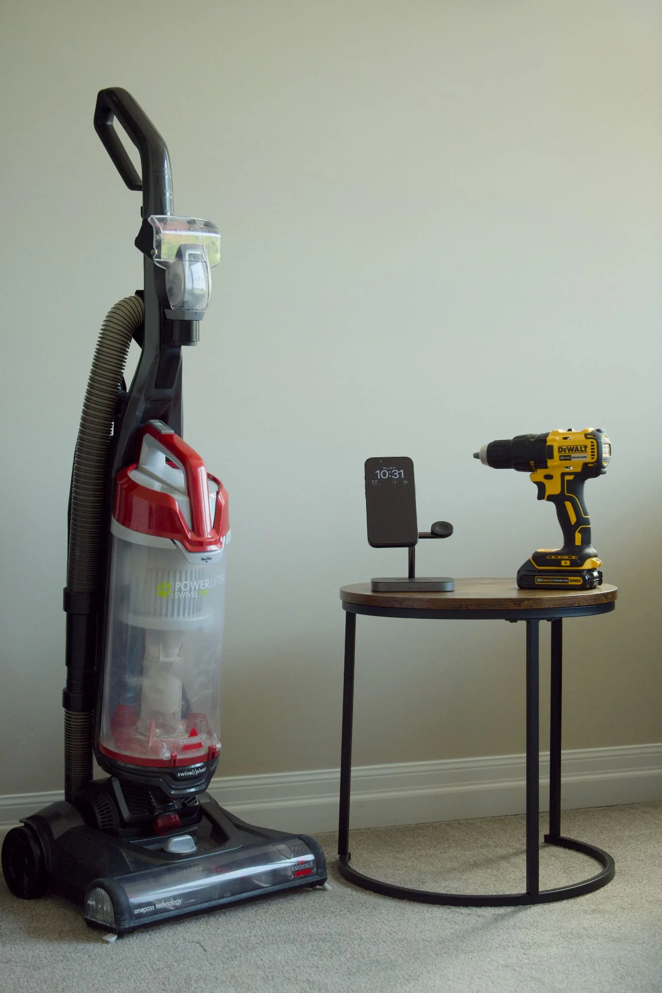 Upright vacuum, wireless phone charger stand, and cordless drill on a small round table.