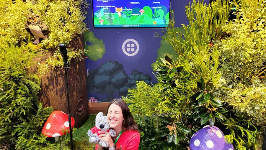 Hugging a stuffed koala in a whimsical forest scene. Twilio logo on the wall surrounded by greenery. Mish is in the middle, cuddling a koala bear that is wearing a red Twilio t-shirt