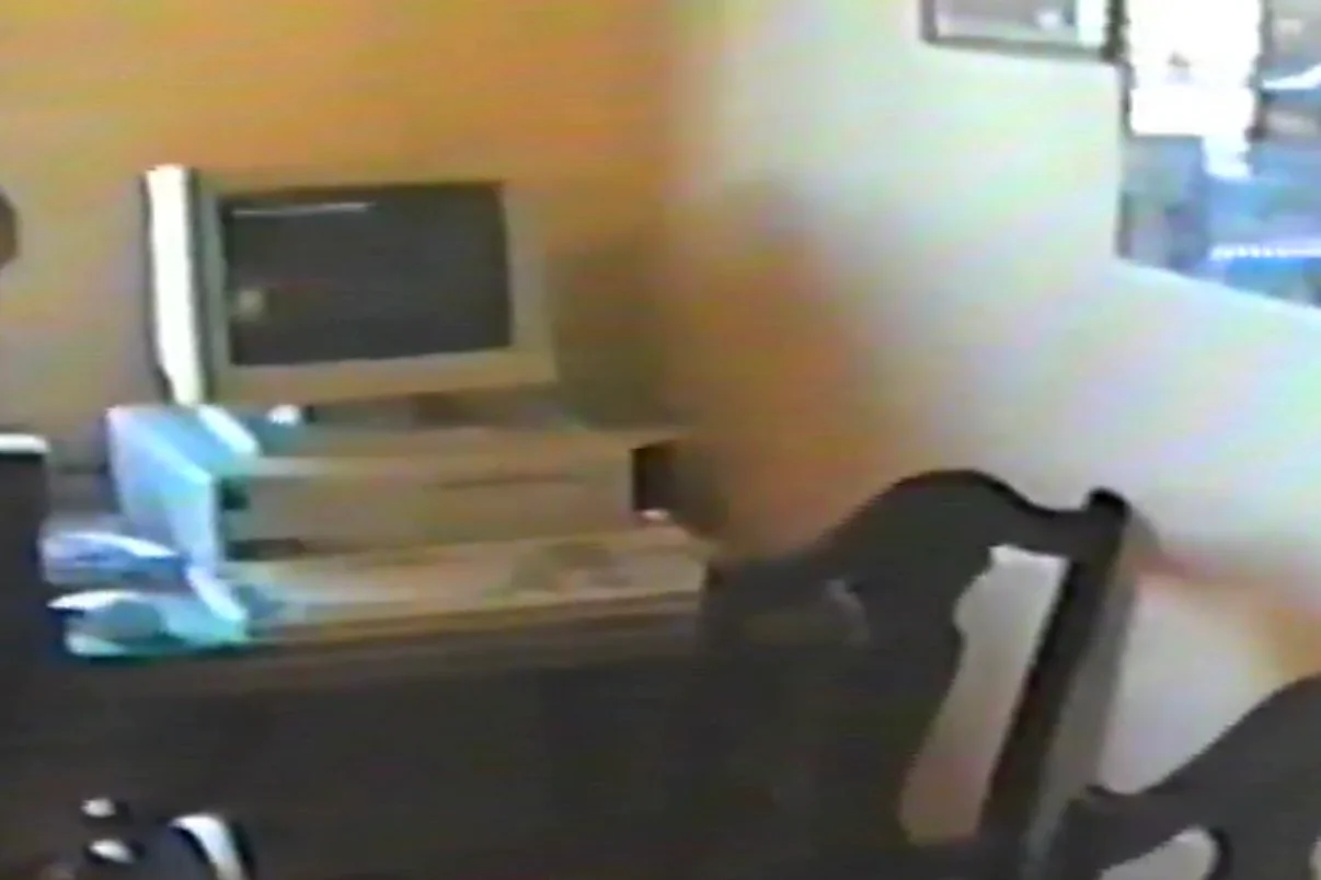 Vintage Home Office Setup with Computer Grainy home video still showing a vintage desktop computer setup from the 1990s, including a CRT monitor and beige tower on a dark wooden desk beside a carved chair.