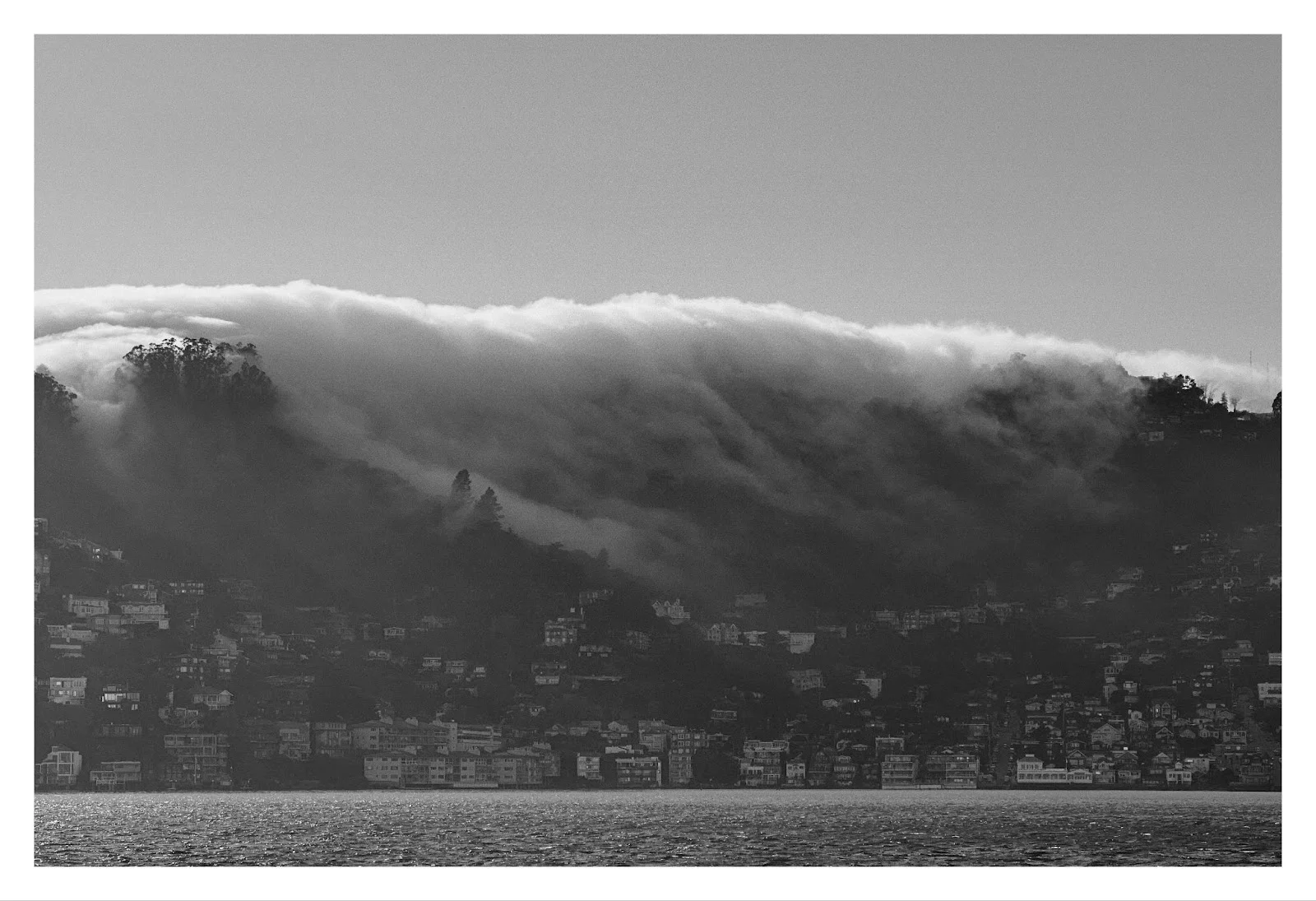 Aerial view of San Francisco skyline with a colorful sunset and the Bay Bridge in the background. Black and white photo of a fog bank rolling over the densely built hillside town of Sausalito by the water, partially obscuring trees and homes as it spills down the slope under a clear sky.