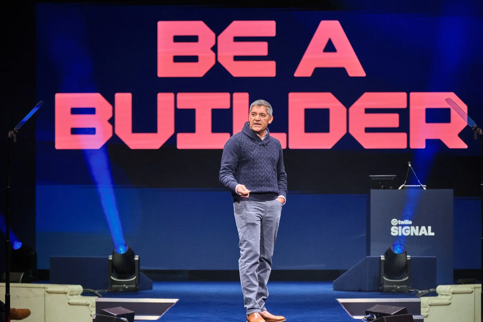Man speaking on stage with BE A BUILDER in large red letters behind him.