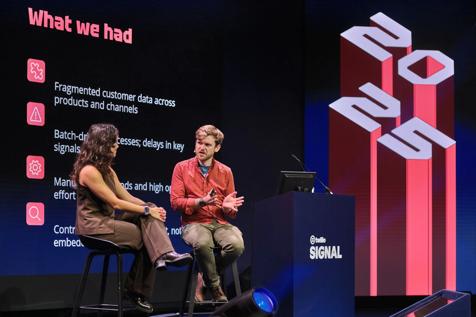 Two presenters on stage discussing What we had with a screen listing fragmented data and high costs at SIGNAL conference.