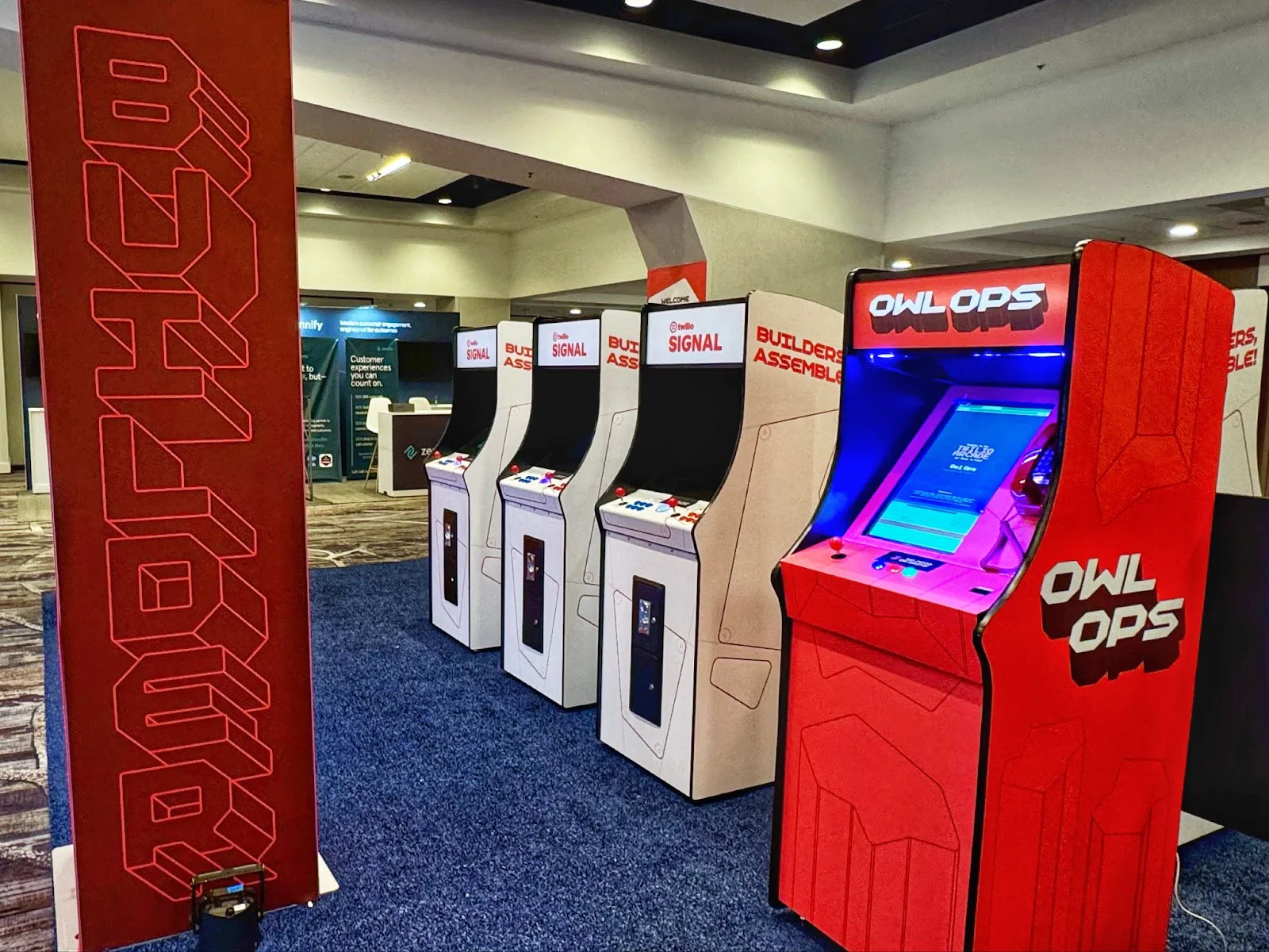 Four arcade machines at a tech expo with screens and 'Builders' and 'Owl Ops' text. Four arcade machines at a tech expo with screens and 'Builders' and 'Owl Ops' text.