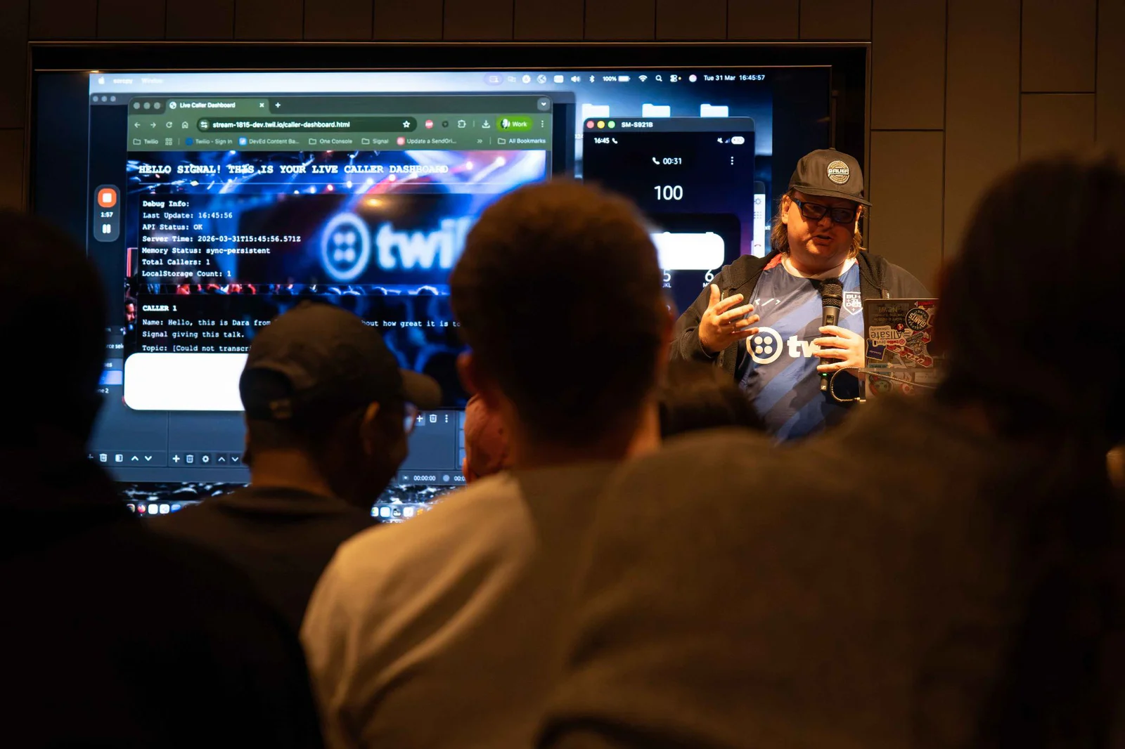 A speaker presents live coding on a screen to an audience at a tech conference.