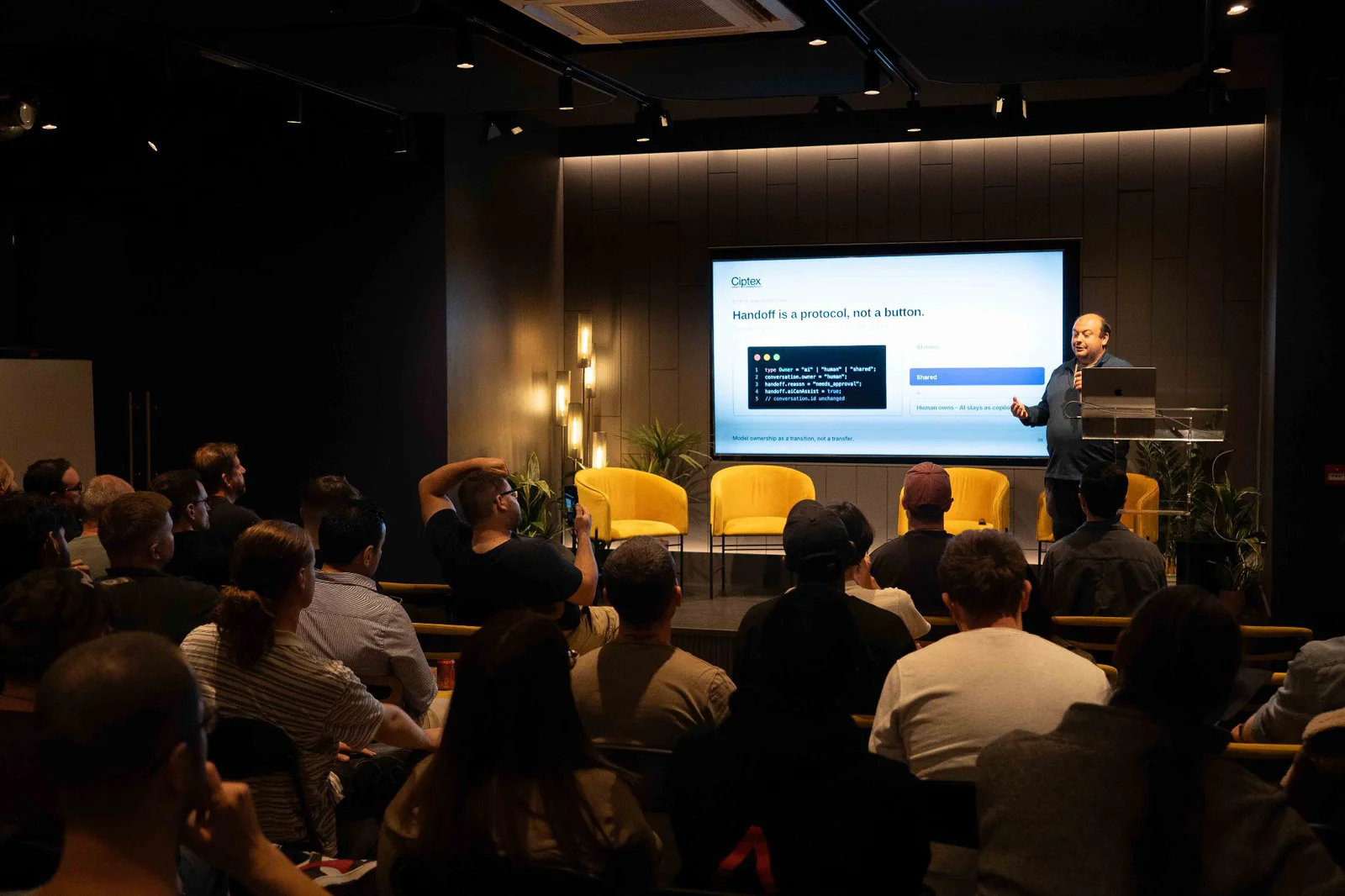 Speaker presenting on Hand-off protocol to an audience at a conference with a large screen behind them.