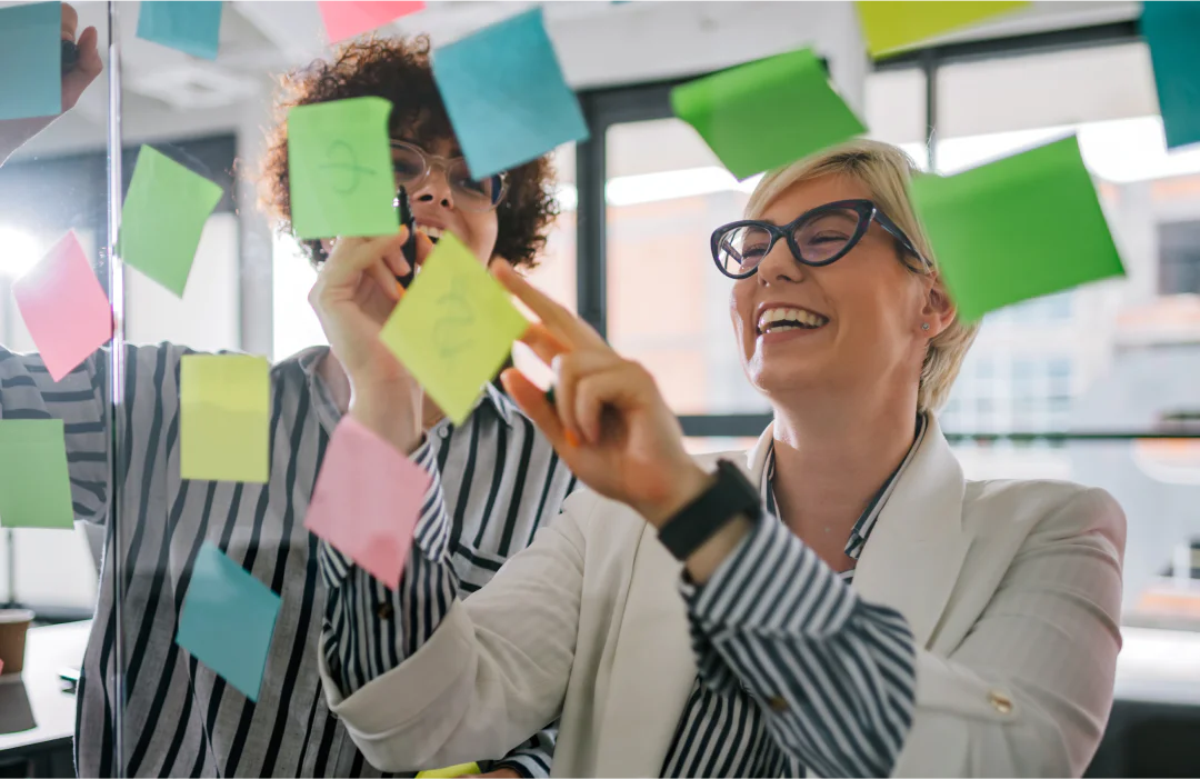 Two colleagues placing colorful sticky notes on a glass wall during a brainstorming session.