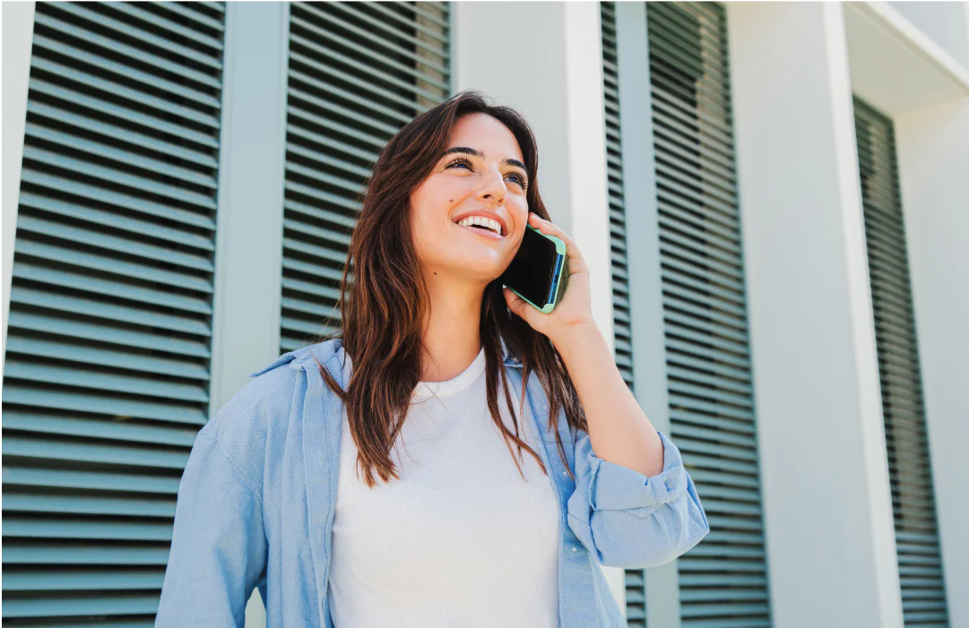 Woman in a blue shirt smiling and talking on a smartphone outside a building with blue shutters.