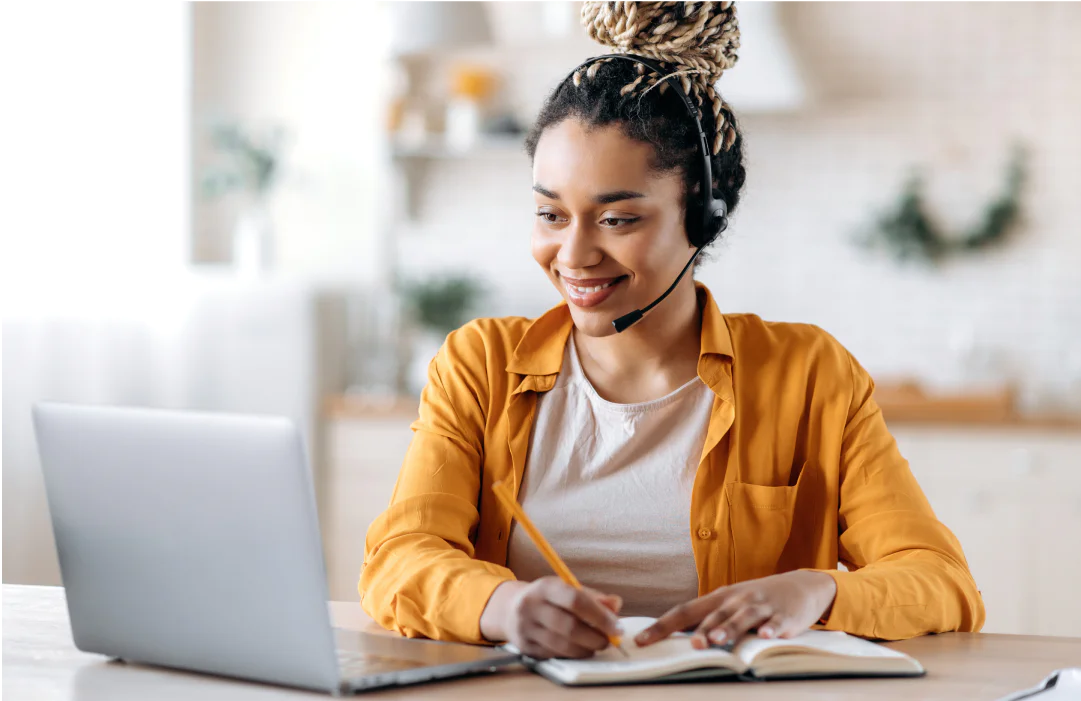 Smiling woman in yellow shirt taking notes while working on laptop at home with a headset on.