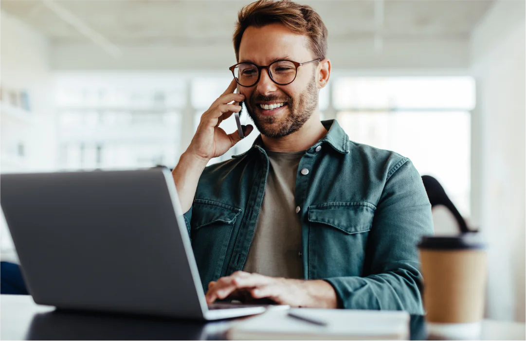 Man wearing glasses talking on phone and using laptop in a modern office