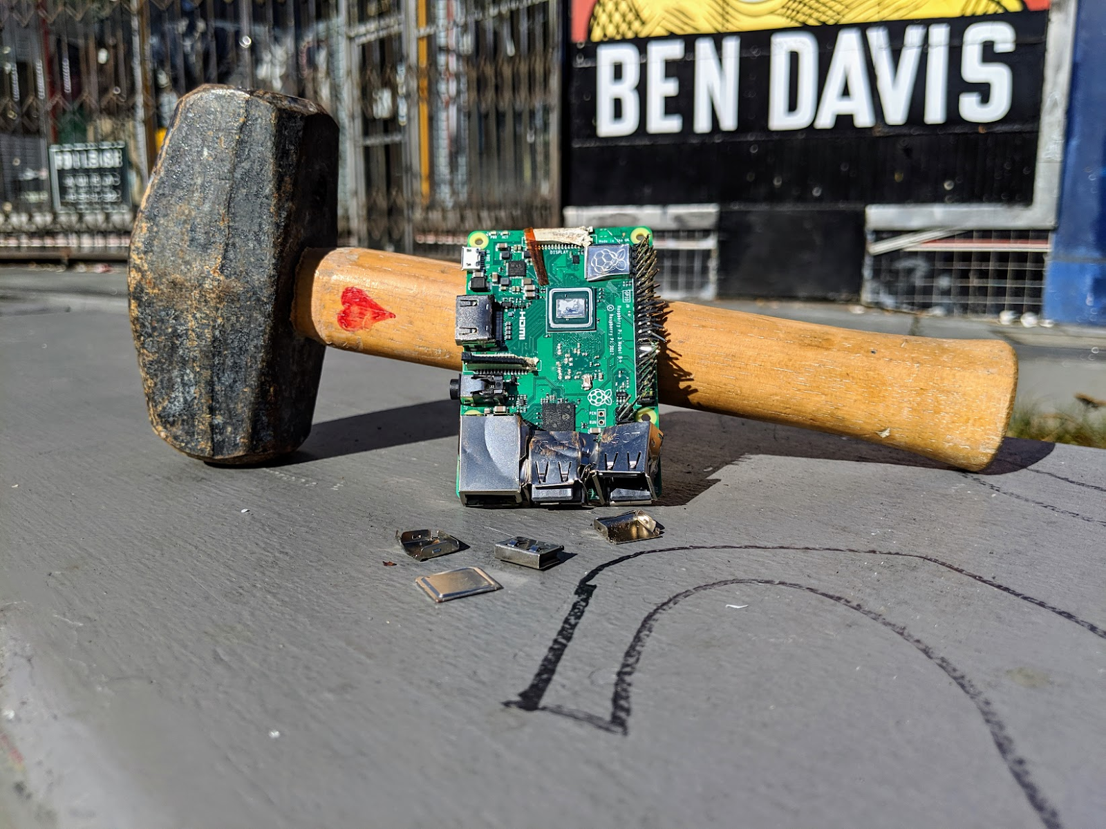A smashed up Raspberry Pi sits propped against a mallet, on top of a concrete barrier. The mallet has a small heart drawn on it.
