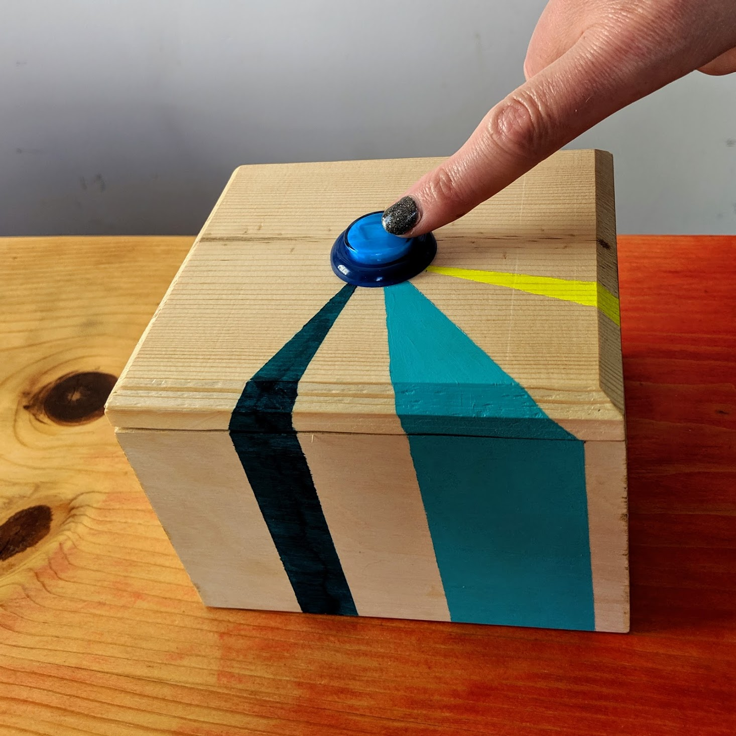 A wooden box with a blue button on the top sits on a table. The box has turquoise, teal, and neon yellow stripes and a finger is pointing at the button.