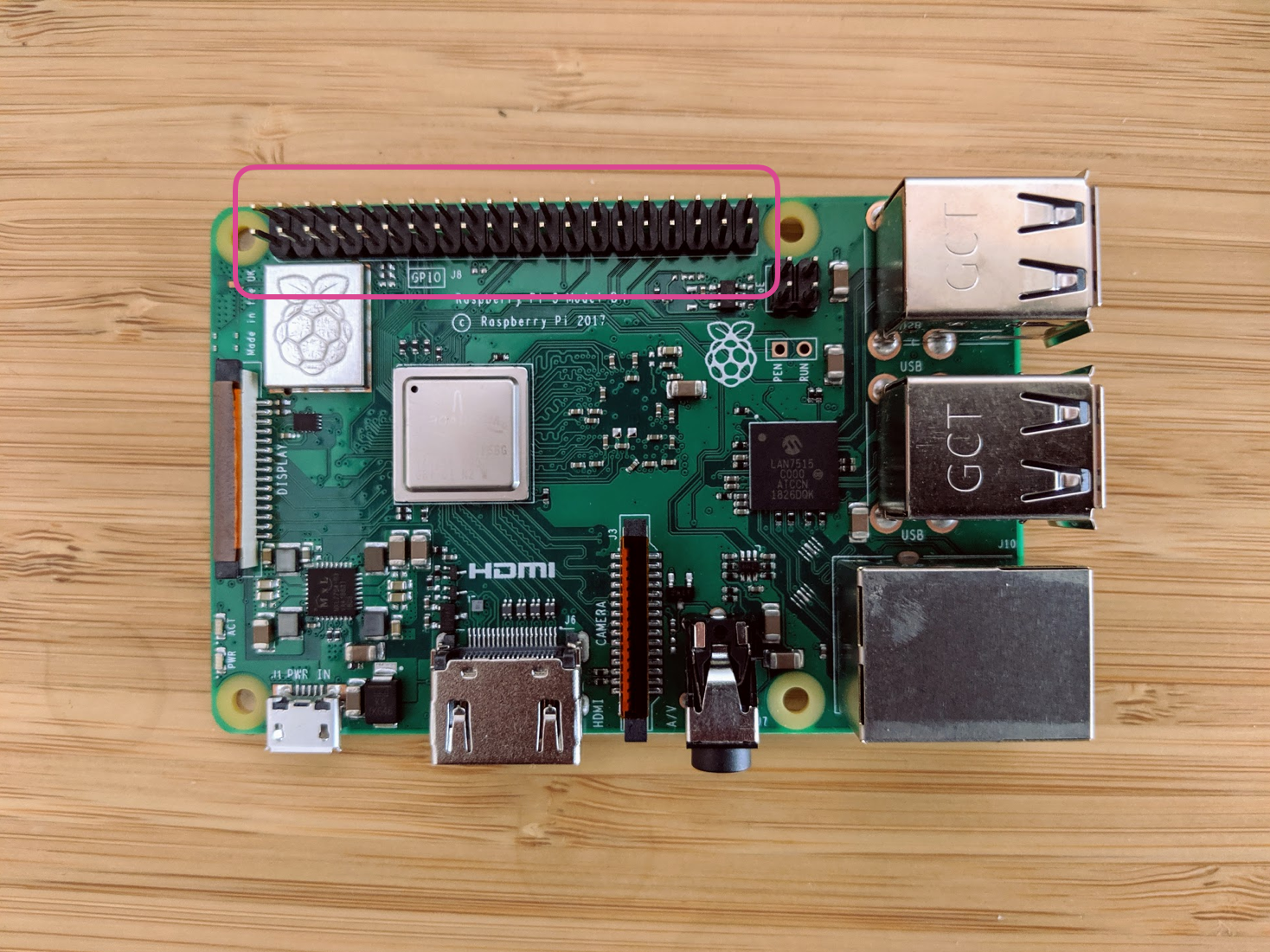 A Raspberry Pi sits on a table. There is a pink circle around the GPIO pins, to indicate their position on the board.