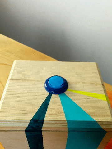 a GIF of a hand with painted nails pressing a button that's attached to a wooden box with turquoise, teal, and neon yellow stripes.