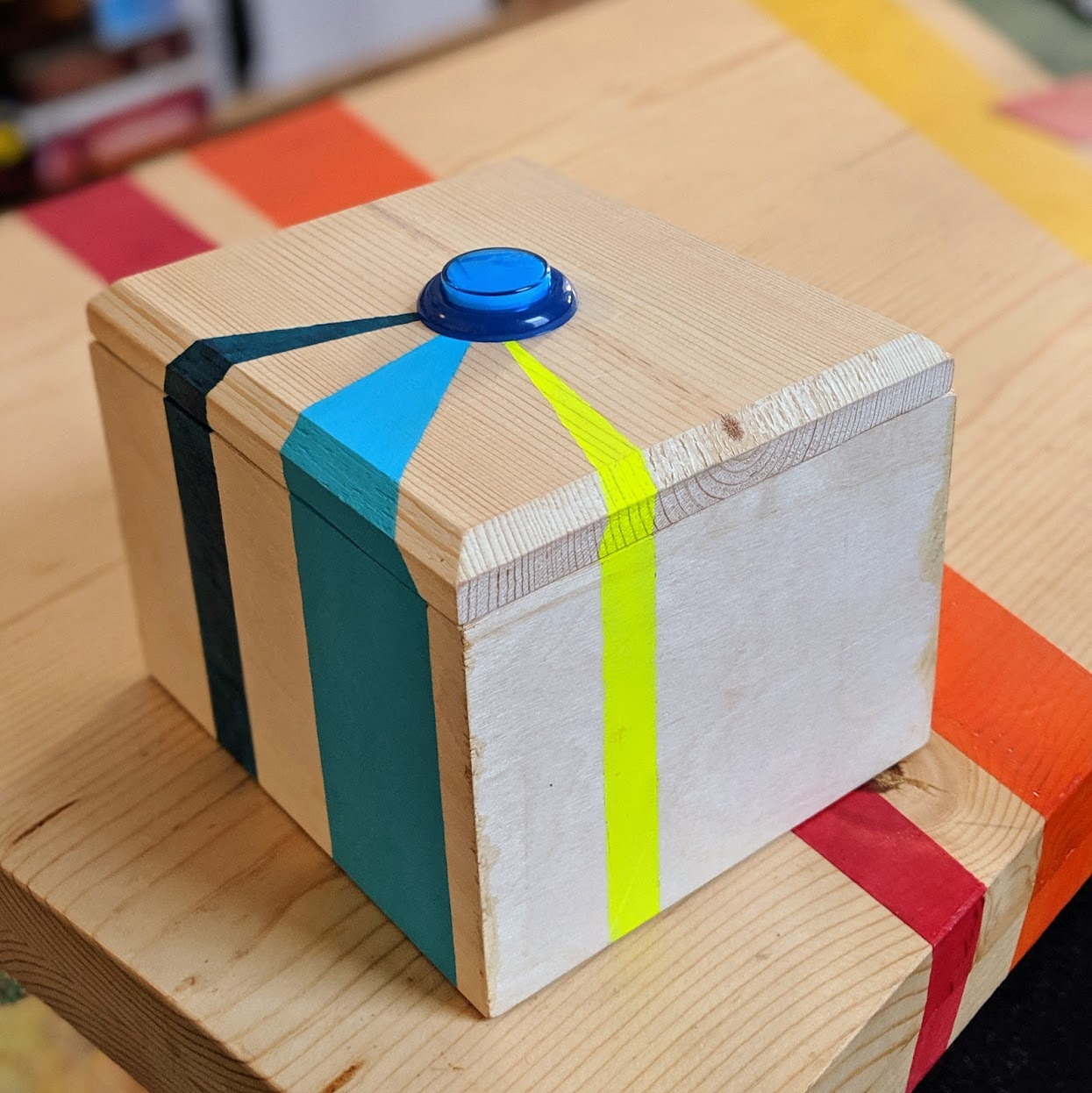 A wooden box with a blue button on the top sits on a table. The box has turquoise, teal, and neon yellow stripes.