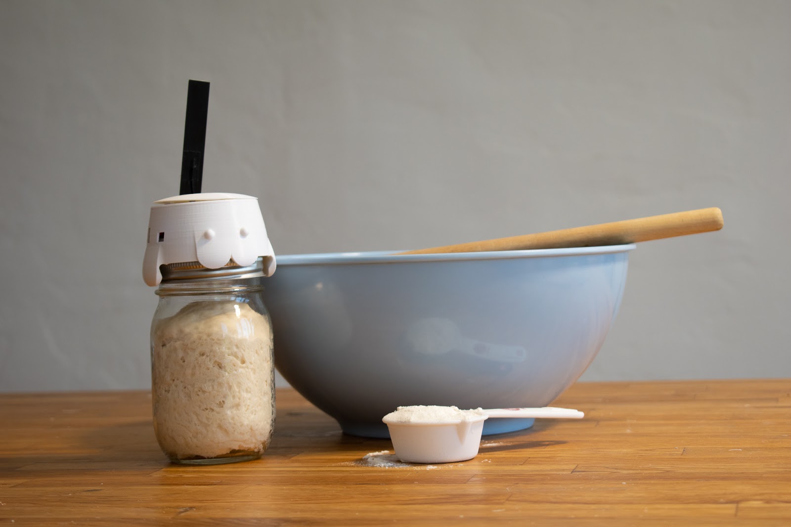 A sourdough starter monitor with a cute face next to a bowl and a small cup of flour