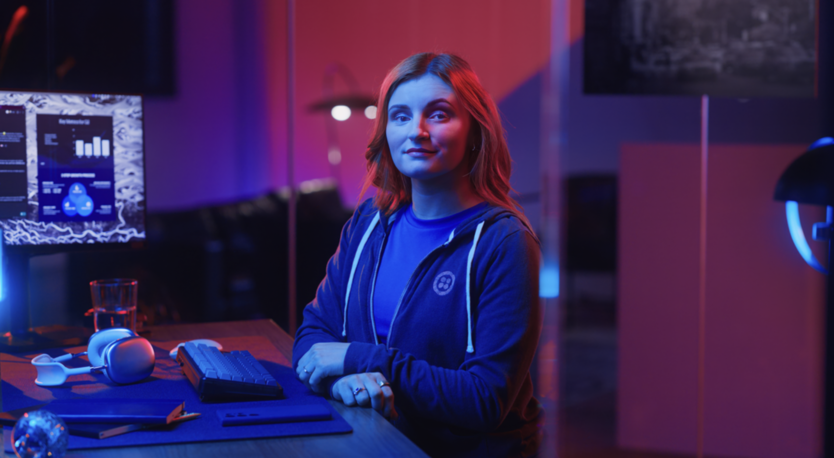 Woman sitting at a desk with computer equipment, in a room lit with blue and pink lights.