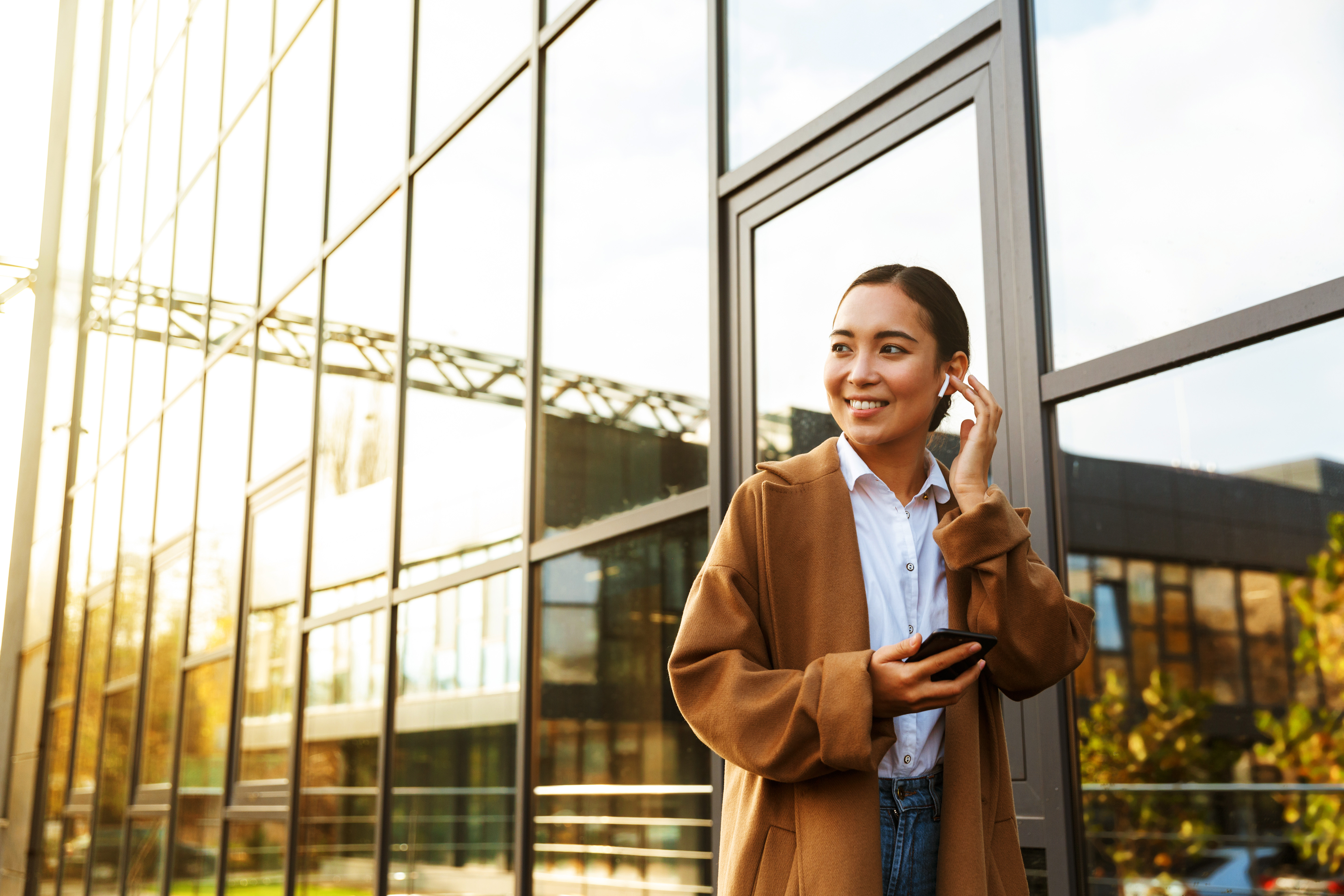 Woman in a brown coat smiling and holding a phone next to a glass building.