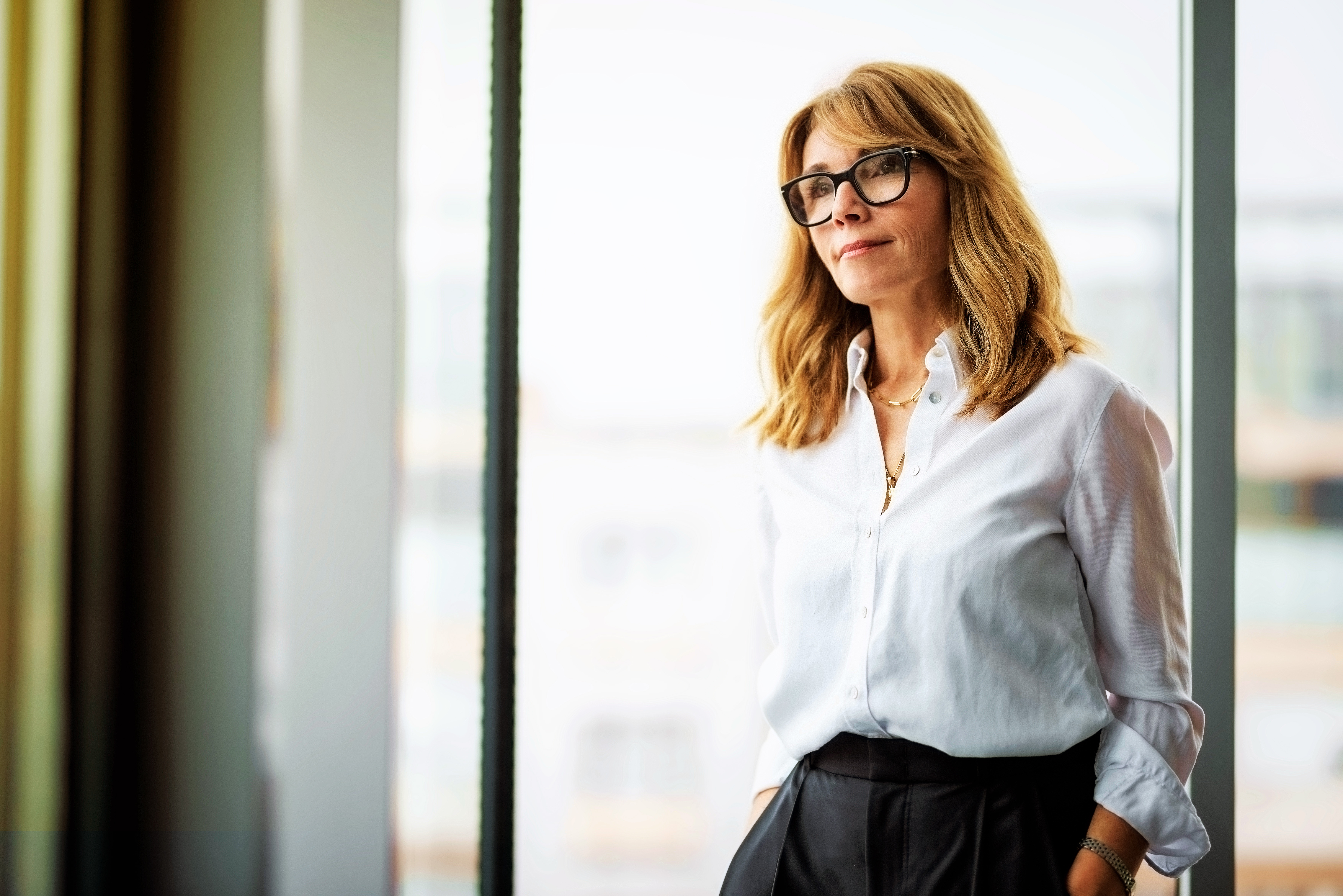 Businesswoman in white shirt and glasses standing by a window, hands in pockets, looking thoughtful.