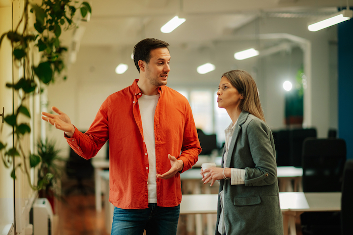 Man in an orange shirt and woman in a blazer talking in an open-plan office.