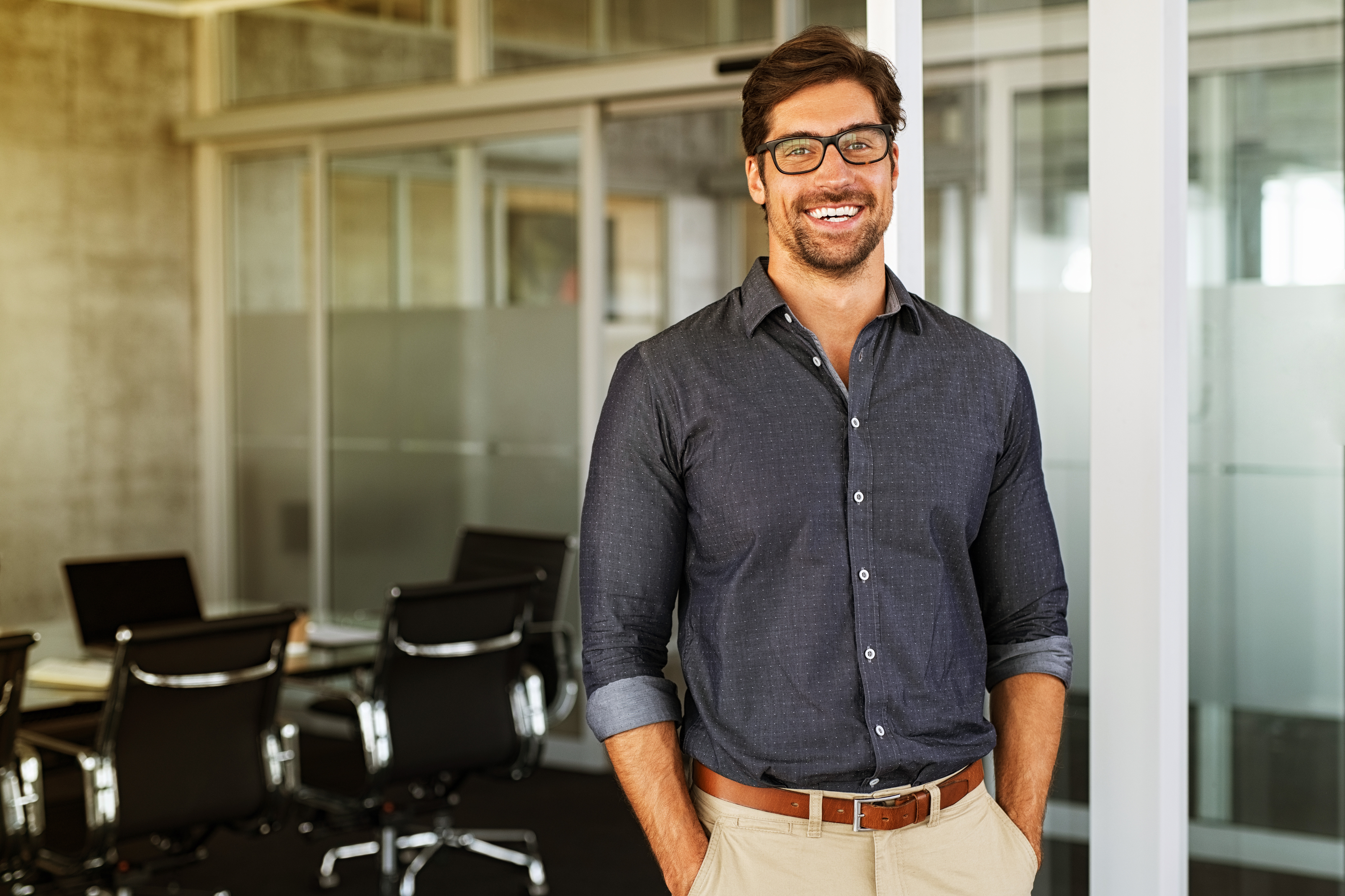 Man with glasses smiling and standing in office with glass walls and chairs in the background.