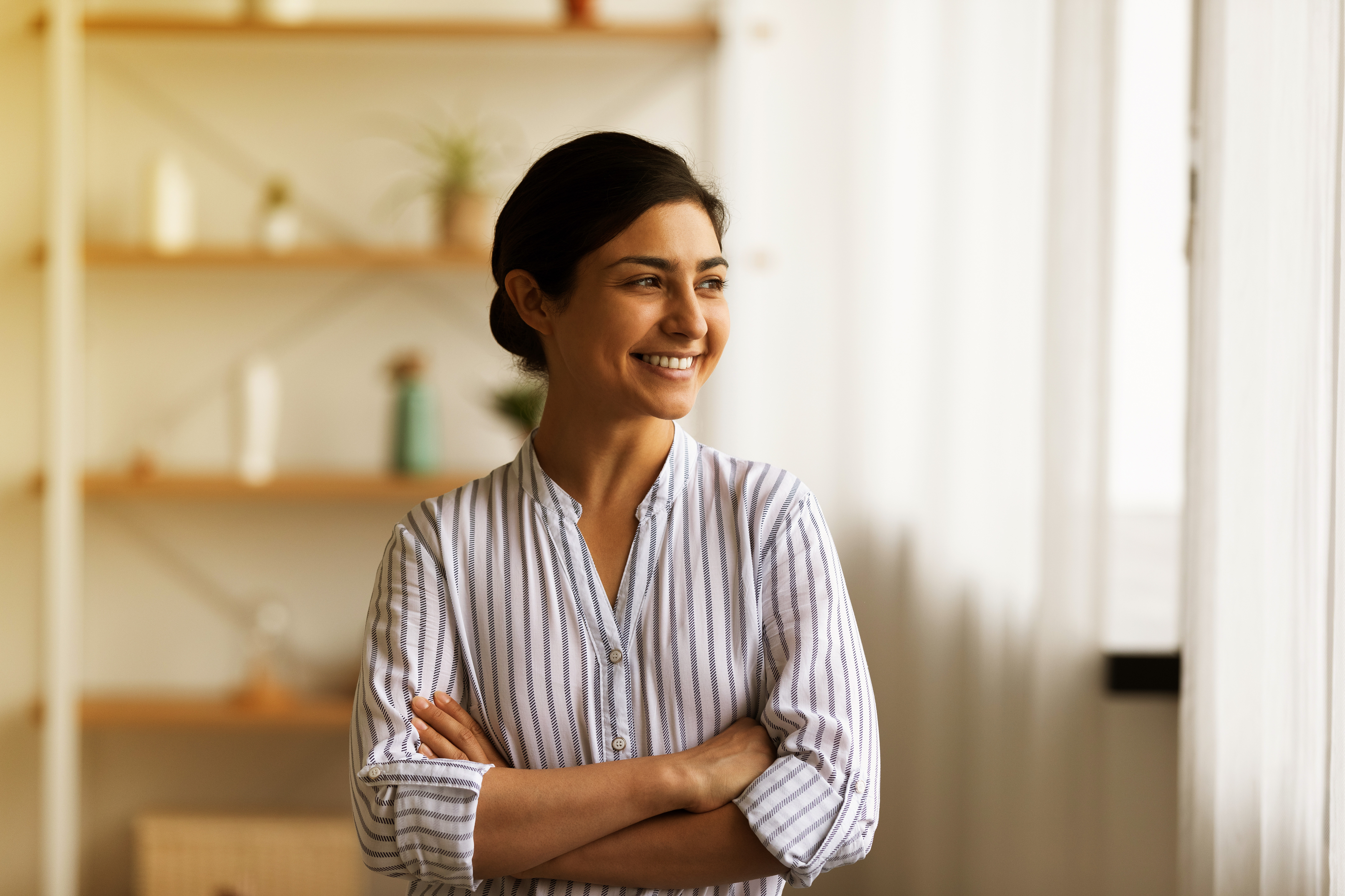 Woman in a striped shirt standing with arms crossed, smiling, with shelves and plants in the background.