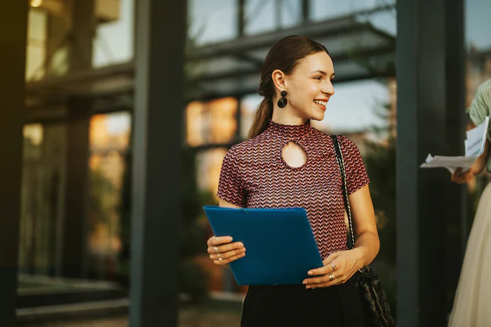 Woman holding a blue folder, smiling, and standing outdoors near a modern building.