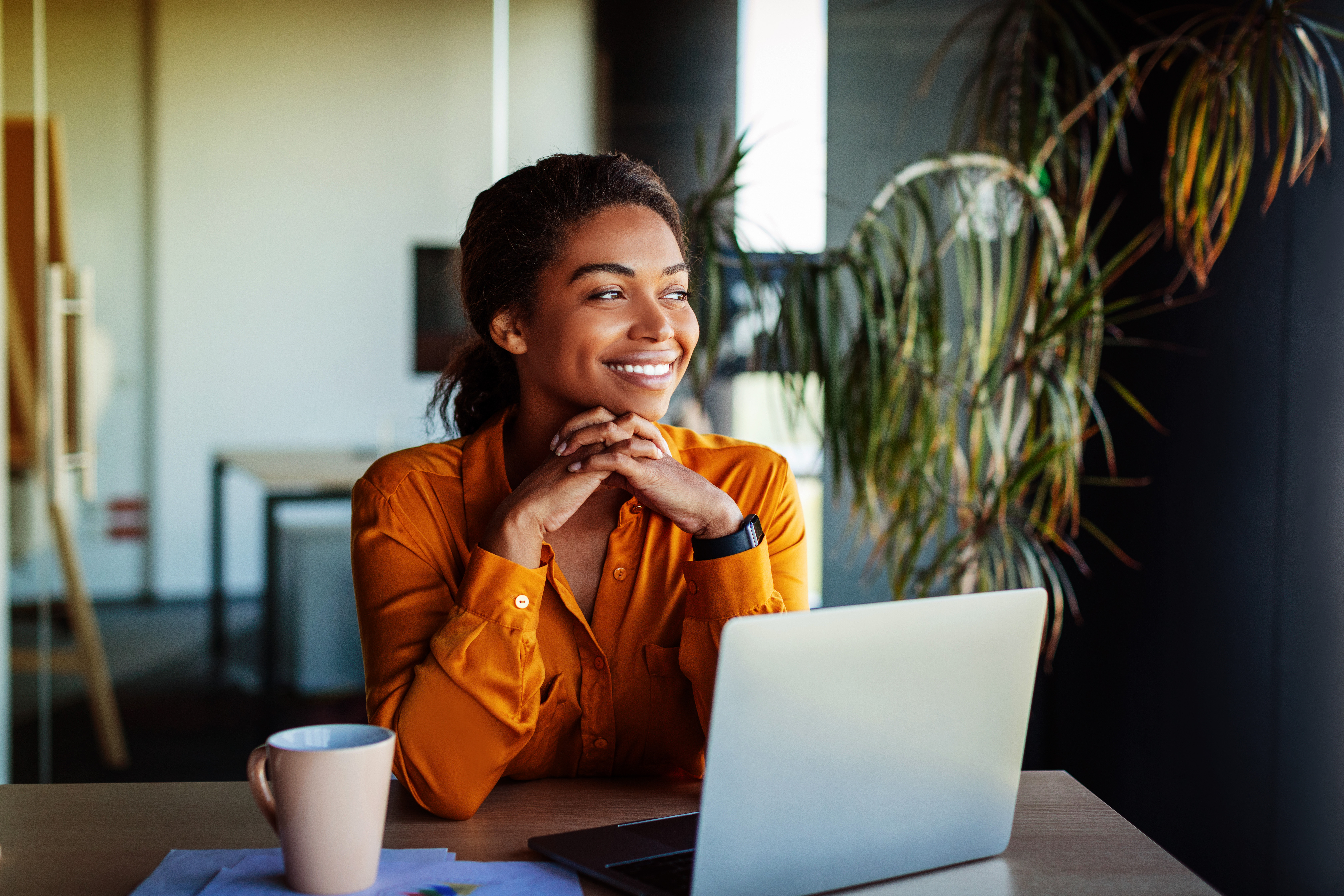 Woman in orange shirt smiling and looking out window, sitting at desk with a laptop and coffee cup